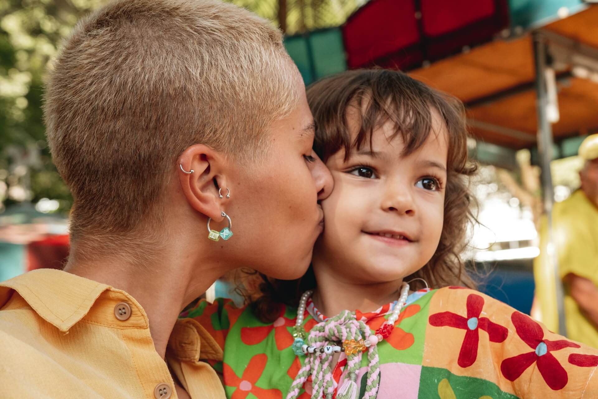A mother with shaved blonde hair kissing her toddler daughter's cheek. they're both dressed in bright clothes.