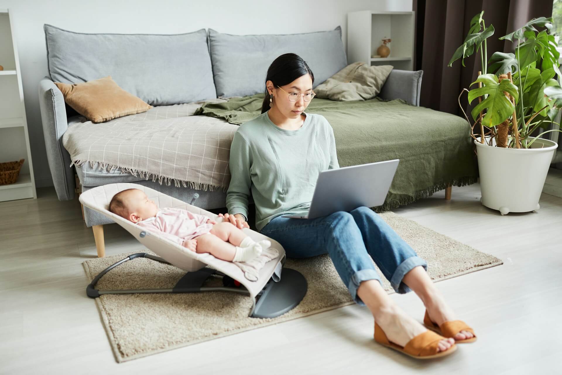 A mother sits on the floor with a laptop on her lap, she is typing with one hand and rocking her baby in a bouncer with the other.